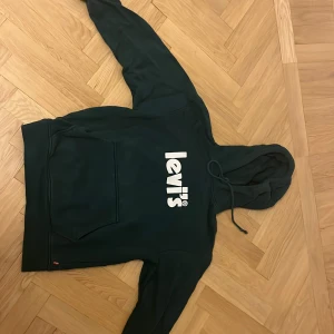 Mörkgrön hoodie från Levi's XS - Mörkgrön hoodie från Levi's i relaxed fit med stor vit logga framtill. Tröjan har huva med snörning och en klassisk känguruficka. Perfekt för dig som gillar en chill och stilren look.