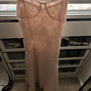 Ljusrosa långklänning med blommönster - Superfin ljusrosa långklänning med subtilt blommönster och markerad byst. Klänningen har en elegant, figurnära passform och är tillverkad i ett tunt, lätt material som ger en romantisk känsla. Perfekt för dig som vill sticka ut med en feminin och stilren look.