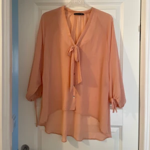 Persikofärgad blus från Atmosphere - Säljer en flowy persikofärgad blus från Atmosphere med v-ringning och knyt framtill. Blusen har trekvartsärmar med knyt vid ärmslut och är tillverkad i ett tunt, lätt material som ger en luftig känsla. Perfekt för en chill och stilren look.
