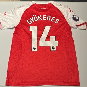 Arsenal Gyökeres 14 matchtröja Adidas - Röd och vit Arsenal fotbollströja från Adidas med Gyökeres och nummer 14 på ryggen. Tröjan har korta ärmar, Premier League-märke på ärmen och klassiskt Arsenal-mönster i tyget. Materialet är lätt och andas, perfekt för match eller träning. Replika, endast handtvätt gäller! Notera defekten på printen på sista bilden! Trycket har släppt lite!