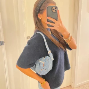 Ljusblå jeans axelväska med nitar - Snygg ljusblå axelväska i jeansmaterial med silverfärgade nitar och dragkedja framtill. Väskan har justerbart axelband och coola detaljer som ger en trendig vibe. Perfekt storlek för det viktigaste och passar till många outfits. Lånade bilder!