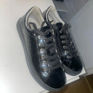 Svarta sneakers från Tommy Hilfiger - Säljer ett par svarta sneakers från Tommy Hilfiger med blank yta och präglat TH-mönster på sidorna. Skorna har svarta snören, dragkedja på sidan och rund tå. Insidan är ljusgrå och bak finns den klassiska Hilfiger-loggan. Perfekt för dig som gillar stilrena och coola detaljer.