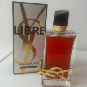 Yves Saint Laurent Libre Le Parfum - YSL Libre Le Parfum med lyxig, rektangulär glasflaska fylld med djup bärnstensfärgad parfym. Flaskan har en elegant svart kork och detaljer i guld, inklusive den ikoniska YSL-loggan. Kommer i stilren vit och guld kartong med svart text.