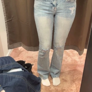 True Religion ljusblå bootcut jeans - Säljer ett par ljusblå bootcut jeans från True Religion med slitna detaljer och röda kontrastsömmar. Jeansen har klassiska fickor med lock och silverfärgade knappar bak samt dragkedja och knappstängning fram. Snygg tvättad look och coola detaljer.