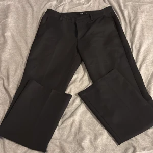 Svarta kostymbyxor från BikBok - Klassiska svarta kostymbyxor från BikBok i storlek 40. Byxorna har raka ben, hällor för bälte och diskreta fickor. Perfekta för en clean och stilren look. Materialet känns mjukt och har en lätt glansig finish.