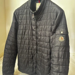 Svart quiltad Moncler pufferjacka - Svart quiltad pufferjacka från Moncler med klassisk logga på ärmen och dragkedja framtill. Jackan har hög krage, ficka med knapp på ärmen och diskreta horisontella sömmar. Perfekt för dig som vill ha en stilren och varm jacka med exklusiv känsla. ( den har ett litet brännmärke som knappt är synligt finns på sista bilden) storlek 4/XL