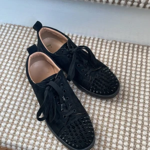 Svarta sneakers med nitar från Christian Louboutin - Riktiga. Skorna kommer med kort och så klart lådan. Snygga svarta sneakers från Christian Louboutin med ikonisk röd sula och nitar på tån. Skorna har snörning och är tillverkade i ett elegant material som ger en lyxig känsla. 