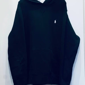 Svart hoodie från Polo Ralph Lauren - Svart hoodie från Polo Ralph Lauren med klassisk vit broderad logga på bröstet. Tröjan har huva, känguruficka och är tillverkad i mjuk bomull. Perfekt för en chill och stilren look.