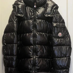 Svart Moncler pufferjacka M - Säljer en svart, glansig pufferjacka från Moncler i storlek M. Jackan har dunfyllning, hög krage, dragkedja framtill och en ikonisk Moncler-logga på ärmen. Perfekt för kalla dagar och har en riktigt fet streetstil.