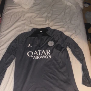 PSG långärmad fotbollströja Jordan - Snygg långärmad PSG fotbollströja i mörkgrått med vita detaljer. Tröjan har Paris Saint-Germain-logga, Jordan-logga och Qatar Airways-tryck på bröstet samt 'Paris' på ärmen. Tillverkad i lätt och ventilerande funktionsmaterial.