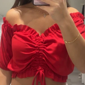 Röd offshoulder croppad topp - Snygg röd offshoulder topp med croppad passform och snörning framtill. Toppen har puffiga korta ärmar och rynkade detaljer längs bröstet och midjan. Perfekt för dig som vill sticka ut med en färgstark och trendig look.