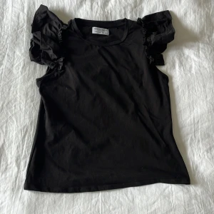 Svart volangtopp från Holly & Whyte - Supersnygg svart topp från Holly & Whyte by Lindex med voluminösa volangärmar. Toppen är ärmlös med rund halsringning och har en clean, rak passform. Perfekt för dig som vill ha en basic topp med en twist. Tillverkad i mjuk bomull. Aldrig använd.