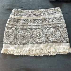 Mönstrad kortkjol med fransar från Maje - Superfin kortkjol från Maje i beige och grått med unikt vävt mönster och dekorativa fransar längs nederkanten. Kjolen har en snygg struktur och känns både trendig och bohemisk. Perfekt att styla med stickad tröja eller blus för en cool look.