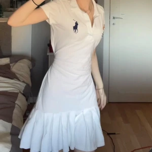 Vit pikéklänning från Ralph Lauren - Vit kort pikéklänning från Ralph Lauren med klassisk krage, korta ärmar och volangkant nertill. Stor broderad logga framtill, tryck och broderier med brittiskt tema samt nummer på ryggen. Tillverkad i mjuk bomull, perfekt för en sportig och stilren look.