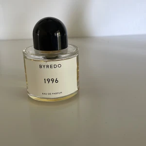 Byredo 1996 Eau de Parfum 50ml - Byredo 1996 Eau de Parfum i 50 ml flaska. Parfymen kommer i en rund, transparent glasflaska med svart kupolformat lock och stilren vit etikett. Doftvätskan är bärnstensfärgad och flaskan är tillverkad i Frankrike.