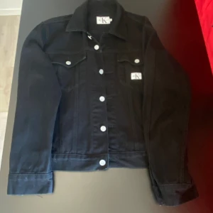 Svart jeansjacka från Calvin Klein - Svart jeansjacka från Calvin Klein Jeans med klassisk krage, silverfärgade knappar och två bröstfickor med lock. Jackan har raka linjer och en stilren look, perfekt för dig som gillar minimalistisk stil. Snygg CK-logga på bröstet.