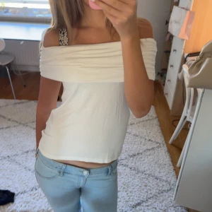Vit offshoulder topp - Säljer en vit offshoulder topp med bred, draperad kant upptill. Toppen är ärmlös och har en enkel, rak passform som sitter snyggt över axlarna. 