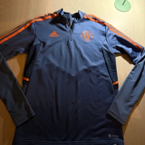 Manchester United långärmad träningströja - Säljer en blå långärmad Manchester United träningströja från Adidas med orange detaljer och klubbmärke på bröstet. Tröjan har dragkedja vid halsen och är tillverkad i funktionsmaterial (AEROREADY) som andas. Perfekt för fotbollsträning eller supporterliv.