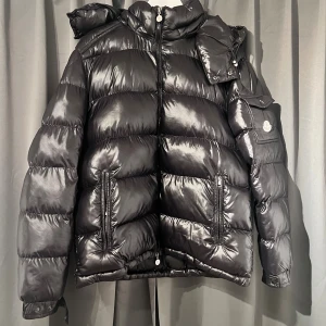 Svart Moncler pufferjacka med huva - Snygg svart pufferjacka från Moncler med glansig finish, vadderad design och avtagbar huva. Jackan har dragkedja framtill, två fickor med dragkedja och en ärmficka med Moncler-logga. Perfekt för kalla dagar och riktigt street.