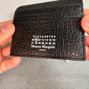 Maison Margiela svart korthållare - Svart korthållare från Maison Margiela i exklusivt präglat skinn. Ikonisk logotyp med siffror framtill och vita sömmar på baksidan. Flera kortfack och stilren design, perfekt för dig som vill ha något både snyggt och praktiskt. Kommer med originalpåse och box. VID NÅGRA ANDRA FRÅGOR SKRIV TILL MIG (PRISET ÄR INTE HACKAT I STEN)
