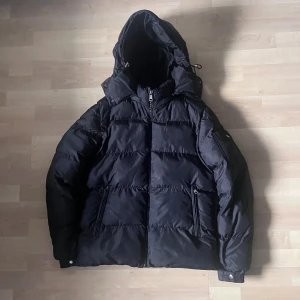 Svart dunjacka från Moncler - Snygg svart dunjacka från Moncler med stor huva och dragkedja framtill. Jackan har två fickor med dragkedja och coolt serietidningsfoder på insidan. Perfekt för kalla dagar och har en klassisk pufferstil som alltid är trendig.