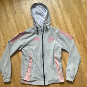 Grå Arsenal vindjacka från Adidas - Snygg grå vindjacka från Adidas med Arsenal-logga och rosa detaljer. Jackan har huva med snörning, hel dragkedja och elastiska muddar. Rosa ränder på ärmarna och paneler vid fickorna ger en sportig vibe. Perfekt för dig som gillar fotboll och streetstyle.