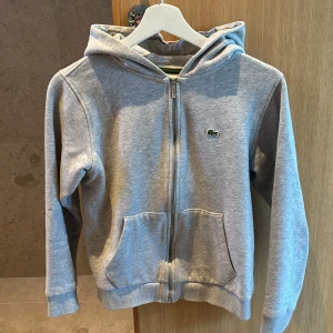 Grå hoodie från Lacoste med dragkedja - Snygg grå hoodie från Lacoste med klassisk krokodillogga på bröstet. Tröjan har huva, långa ärmar, två fickor fram och hel dragkedja.