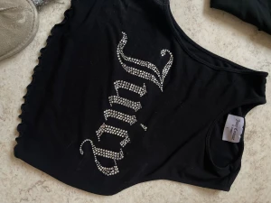 Svart Juicy Couture linne med strass - Säljer ett svart linne från Juicy Couture med glittrande strass-stenar som bildar texten 'Juicy' på framsidan. Linnet har vågiga kanter och ärmlös design, perfekt för en trendig och cool look.