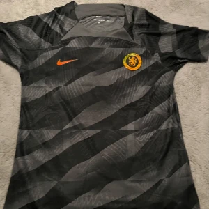 Chelsea FC svart kortärmad fotbollströja - Snygg Chelsea FC fotbollströja från Nike i svart och grått med ett unikt mönster. Kortärmad modell med orange Nike-logga och Chelsea-emblem i orange och svart på bröstet. Tillverkad i lätt och ventilerande material, perfekt för match eller träning.