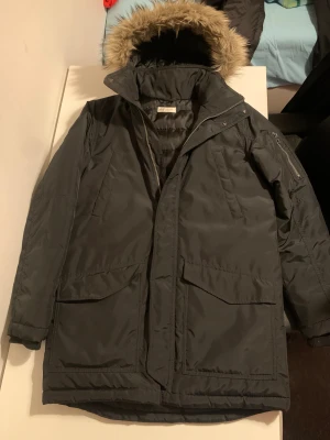 Svart parkas från H&M med fuskpäls - Svart parkas från H&M med huva och fuskpäls, perfekt för kalla dagar. Jackan har flera fickor, dragkedja och knappar framtill samt quiltad insida för extra värme. Materialet är syntet och polyester, och modellen är lång med normal passform.