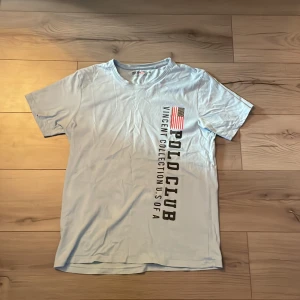 Ljusblå Polo Club t-shirt i bomull - Snygg ljusblå t-shirt från Polo Club Saint Vincent med tryck på bröstet och amerikansk flagga. Modellen är klassisk med rund hals och korta ärmar. Tillverkad i 100% mjuk bomull, perfekt för en chill och stilren look.