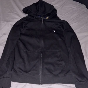 Svart hoodie från Polo Ralph Lauren XL - Svart hoodie från Polo Ralph Lauren i storlek XL. Klassisk design med huva, dragkedja framtill och två fickor. Liten vit logga broderad på bröstet. Perfekt för chill dagar och enkel att matcha med jeans eller joggers.