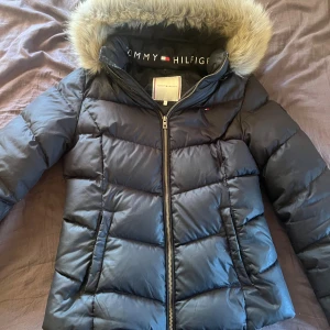 Svart pufferjacka Tommy Hilfiger - Svart pufferjacka från Tommy Hilfiger med snygg pälsimitation på huvan och logotyp på insidan. Jackan har quiltad design, dragkedja framtill och två fickor. Perfekt för kalla dagar och har en modern, stilren look.