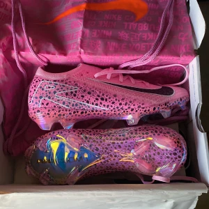 Nike rosa fotbollsskor med holografisk sula - Säljer ett par sjukt snygga fotbollsskor från Nike i rosa med svarta och blå prickar. Skorna har snörning och en holografisk sula som verkligen sticker ut på planen. Lätta och smidiga med rund tå och platt sula, perfekta för dig som vill ha max stil och prestanda.