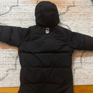 Sail racing jacka - Svart dunjacka från Sail Racing med huva och coola detaljer som Sail Antarctica Expedition-märke på ärmen. Jackan har flera fickor med dragkedja och orange zip-pullers, samt Sail Racing-logga på bröstet. Perfekt för kalla vinterdagar och riktigt snygg stil.