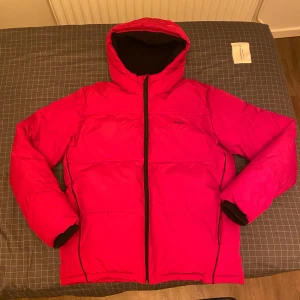 Röd pufferjacka från BOSS med huva - Snygg röd pufferjacka från BOSS med stor logga på ryggen och mindre logga på bröstet. Jackan har huva, svarta detaljer och hel dragkedja framtill. Insidan är svart med BOSS-mönster och jackan är fodrad för extra värme. Perfekt för kalla dagar. Köpt från Zalando och nypris ligger på 2200kr. Bara att skriva vid frågor och priset går såklart att diskutera!