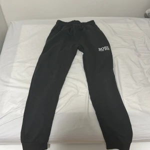 Svarta mjukisbyxor från Hugo Boss - Olivgröna mjukisbyxor från Hugo Boss med broderad logga på vänster lår. Byxorna har elastisk midja, ribbade muddar vid benslut och en praktisk ficka med dragkedja på baksidan. Perfekta för chill eller streetwear-stil.