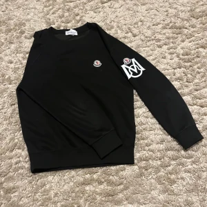 Svart sweatshirt från Moncler med logga - Svart sweatshirt från Moncler med rund hals och ribbade muddar. På bröstet sitter Moncler-loggan och på ena ärmen finns en stor vit tryckt Moncler-symbol. Tröjan har lång ärm och är gjord i mjuk bomull som är skön att ha på sig.