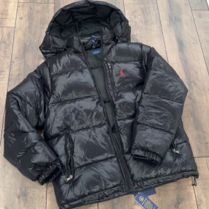 Ralph Lauren vinterjacka  - Svart pufferjacka från Polo Ralph Lauren med huva, dragkedja och två sidofickor. Jackan har en röd Polo-logga på bröstet och justerbar nederkant med snörning. Materialet är glansigt och känns riktigt chill för kalla dagar.