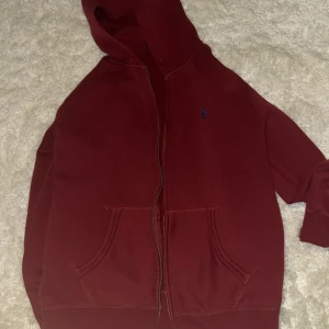 Vinröd hoodie från Polo Ralph Lauren - Säljer en vinröd hoodie från Polo Ralph Lauren med dragkedja och huva. Tröjan har två stora fickor framtill och ett litet blått Polo-logo broderat på bröstet. Mjuk insida i fleece och klassisk passform. 1200kr