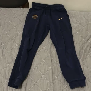 PSG x Nike mörkblå mjukisbyxor - Mörkblå mjukisbyxor från Nike i samarbete med PSG. Byxorna har guldfärgade detaljer, PSG-logga och Nike Swoosh framtill, samt snörning i midjan och muddar vid bensluten. Perfekta för chill eller sport. Materialet är mjukt och bekvämt.