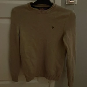 Beige stickad tröja från Morris - Snygg beige stickad tröja från Morris med rund hals och diskret logga broderad på bröstet. Tröjan har långa ärmar och ribbade muddar vid hals, ärmslut och nederkant. Perfekt för dig som gillar klassisk och stilren look.