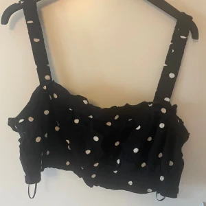 Svart prickig croppad topp H&M - Svart croppad topp från H&M med breda axelband och vita prickar. Toppen har en lätt rynkad struktur och volangdetaljer längs kanterna. Perfekt för dig som gillar en lekfull och trendig stil.