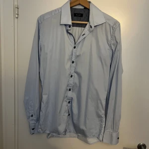 Ljusblå skjorta från Sand Copenhagen - Snygg ljusblå skjorta från Sand Copenhagen i storlek 41. Skjortan har klassisk krage, svarta knappar och lång ärm. Insidan av kragen och knappslån har ett prickigt mönster i vitt och marinblått som ger en cool kontrast. Perfekt för dig som gillar stilrena detaljer. Aldrig använd, perfekt till sommaren, linne, skriv vid funderingar 