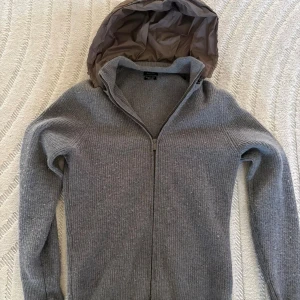 Grå stickad jacka med huva från Massimo Dutti - Snygg grå stickad jacka från Massimo Dutti med dragkedja och quiltad huva i mörkare ton. Jackan är ribbstickad och har långärmade ärmar. Materialet är exklusiv ull som ger en mjuk och varm känsla. Perfekt för dig som gillar stilren och chill look.