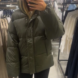 Grön pufferjacka med hög krage - Säljer en mörkgrön pufferjacka med hög krage och stora, quiltade partier. Jackan har långa ärmar och stängs med både dragkedja och knappar framtill. Perfekt för kalla dagar och har en oversized passform som ger en cool och avslappnad look. Ny pris 699; Andvänd en gång så är som ny utan lapp med inga defekter därav priset 