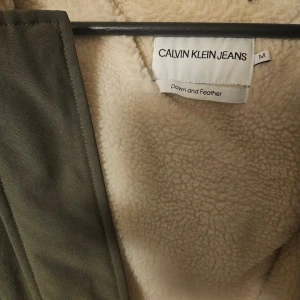 Olivgrön parkas från Calvin Klein Jeans - Parkas i olivgrönt från Calvin Klein Jeans med fuskpäls på huvan och mjukt teddyfoder. Jackan har stora fickor, dragkedja och knappar framtill samt ribbade muddar vid ärmslut. Perfekt för kalla dagar med sin varma dun- och fjäderfyllning.
