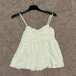 Vit volangig linnetopp med smala axelband - Supersöt vit linnetopp med smala justerbara axelband och volangiga detaljer. Perfekt för varma dagar. Strl Xs