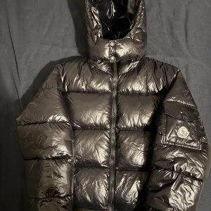 Svart Moncler pufferjacka med huva - Säljer en svart pufferjacka från Moncler i storlek S. Jackan har en glansig finish, huva och dragkedja framtill. Klassisk Moncler-logga på ärmen och coolt serietryck på insidan. Perfekt för dig som vill ha en varm och snygg jacka med street vibe.