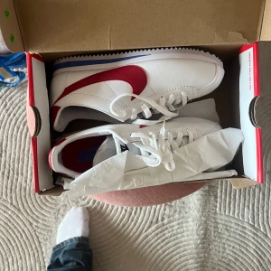 Nike Classic Cortez Leather vit/röd - Nike Classic Cortez Leather sneakers i vitt med röd swoosh och blå detalj på sulan. Skorna har snörning, platt sula och rund tå. Tillverkade i skinn med klassisk retrostil och ikonisk design. Perfekt för dig som gillar sportig och clean look.